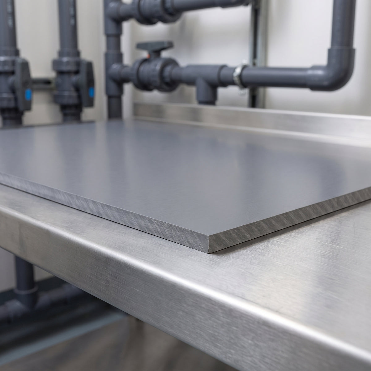 Grey PVC sheet on a stainless steel bench in a chemical processing plant, showing its machined cut edge, with grey PVC-U pipework and valves behind